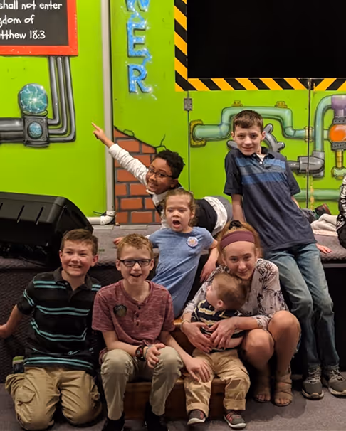 Group of seven children smiling and sitting together indoors against a colorful green wall with decorative pipes and playful designs.