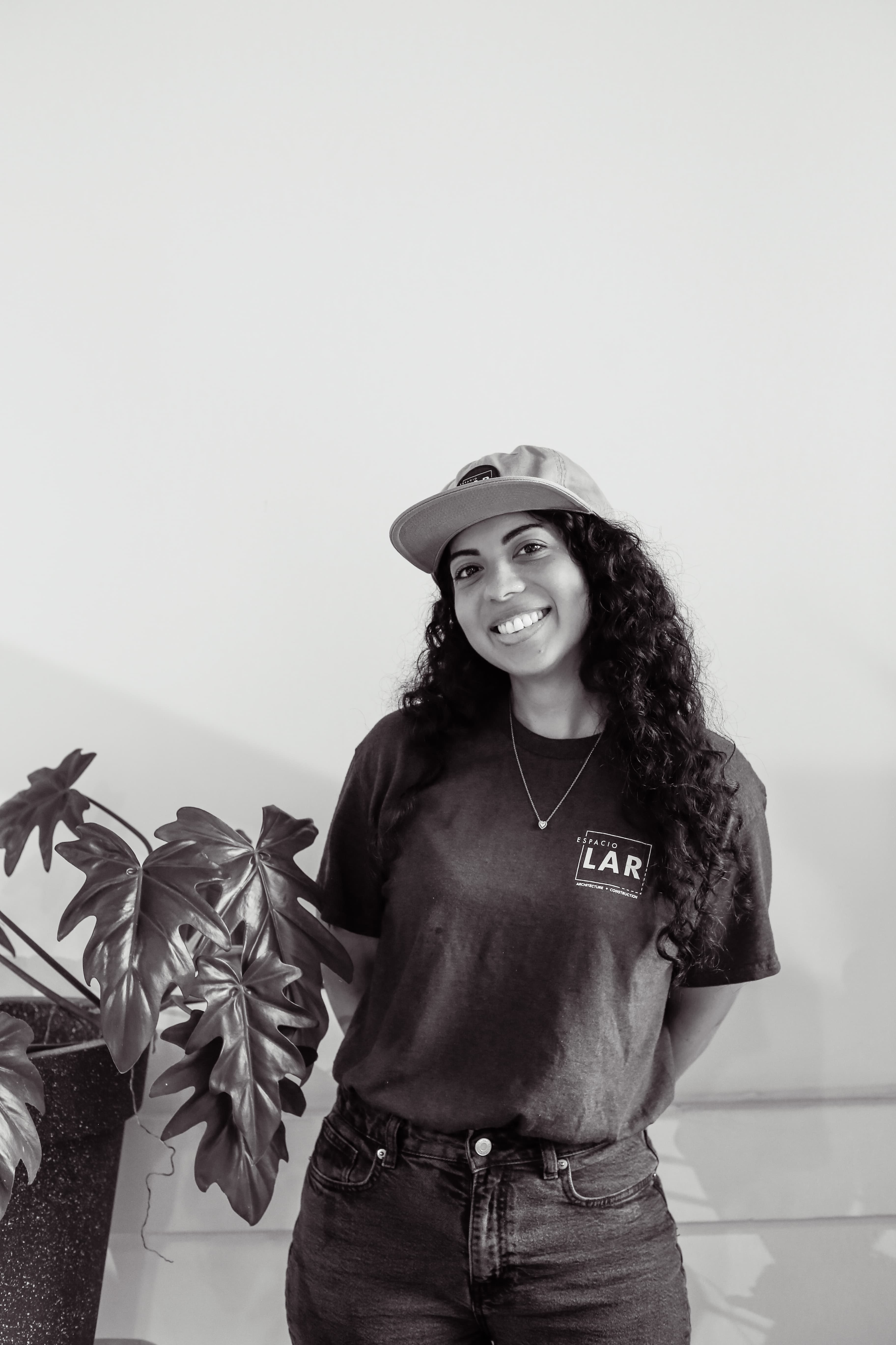 Smiling woman with long curly hair wearing a cap and Espacio LAR t-shirt standing next to a large potted plant.