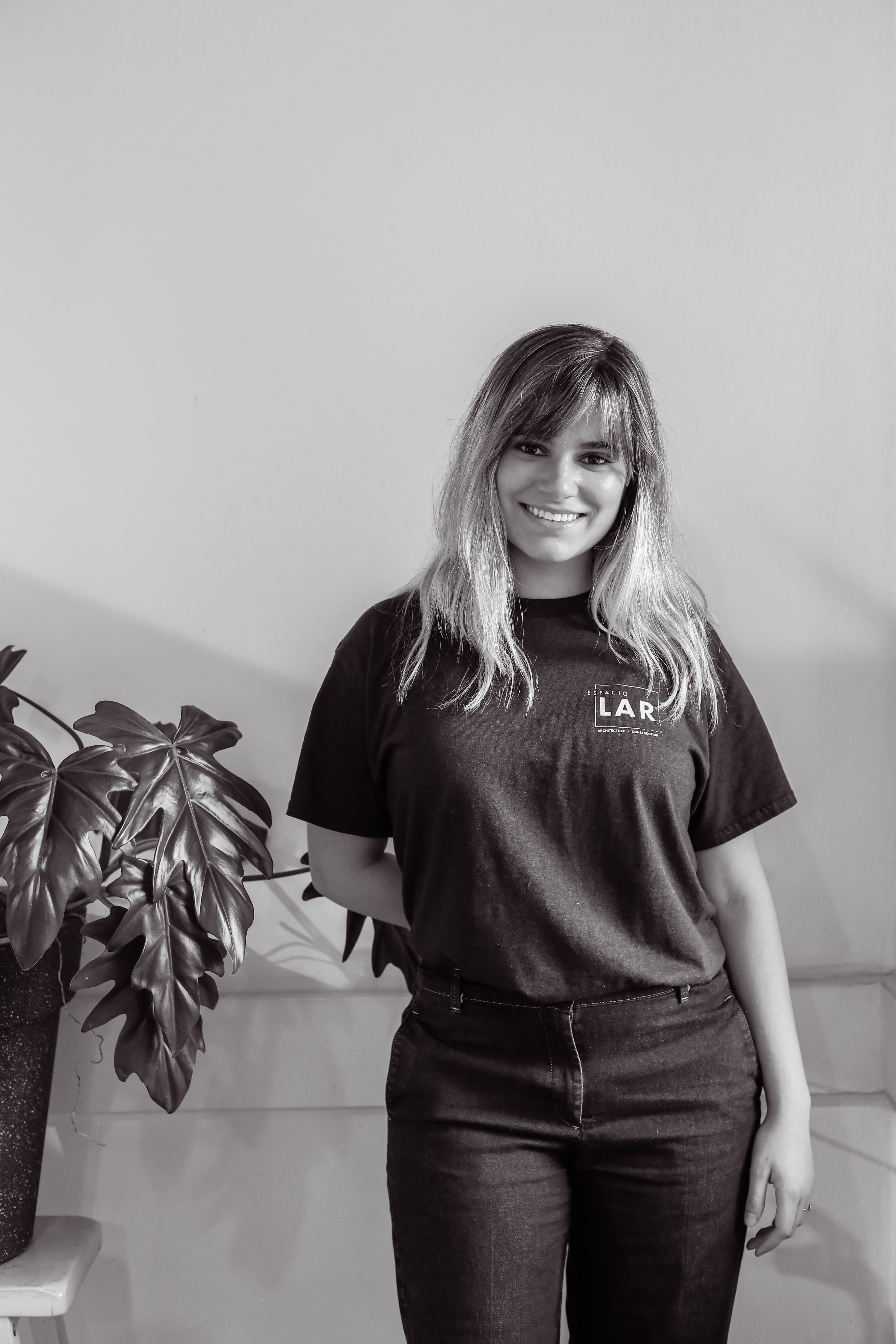 Smiling woman with long hair wearing a black Espacio LAR t-shirt standing next to a large leafy plant.