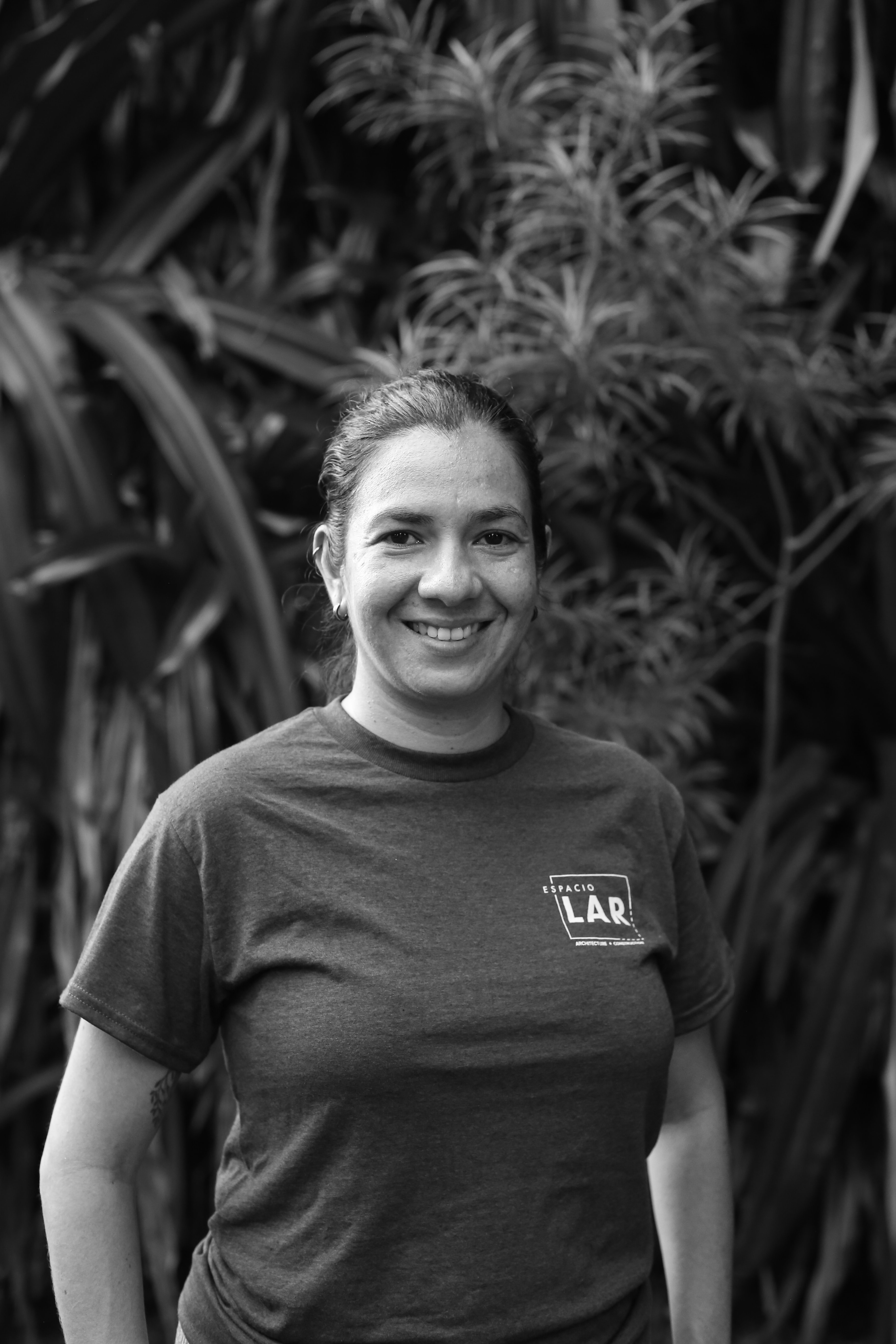 Smiling woman wearing a dark blue Espacio LAR t-shirt standing outdoors in front of green leafy plants.