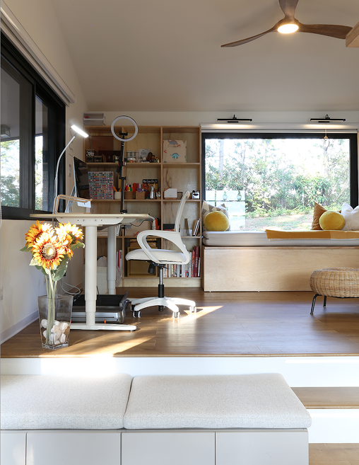 Bright modern home office with white desk, office chair, bookshelf, large window seat with yellow cushions, and a vase of sunflowers.
