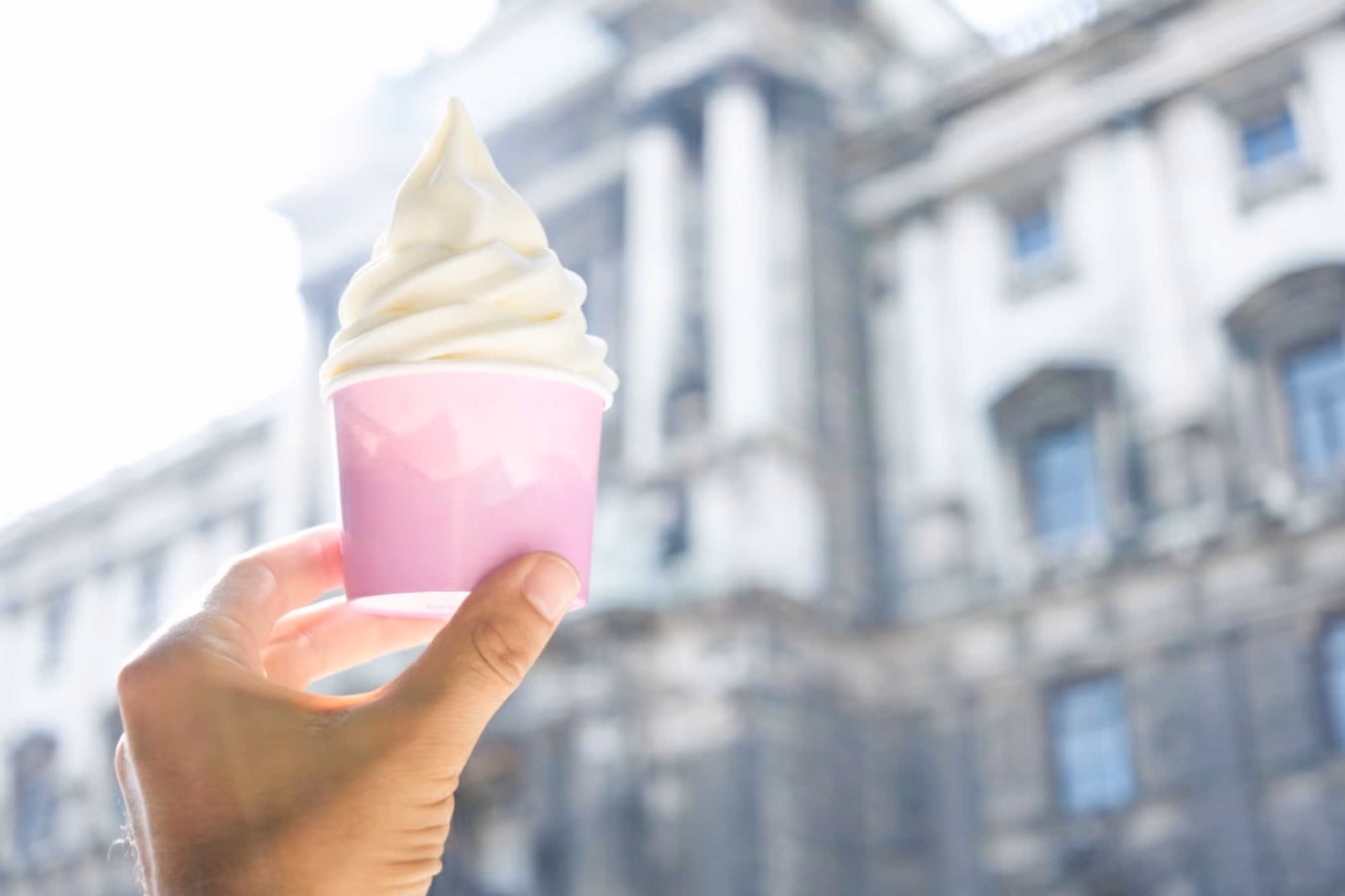 Hand holding a pink cup of soft-serve vanilla ice cream with a blurred historic building in the background.