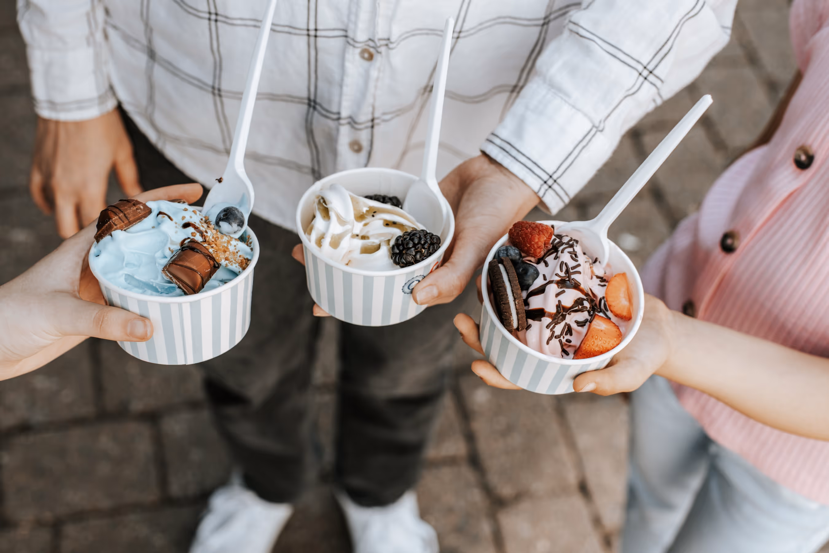 Three people holding cups of frozen yogurt topped with fruits, chocolate, and candy pieces.