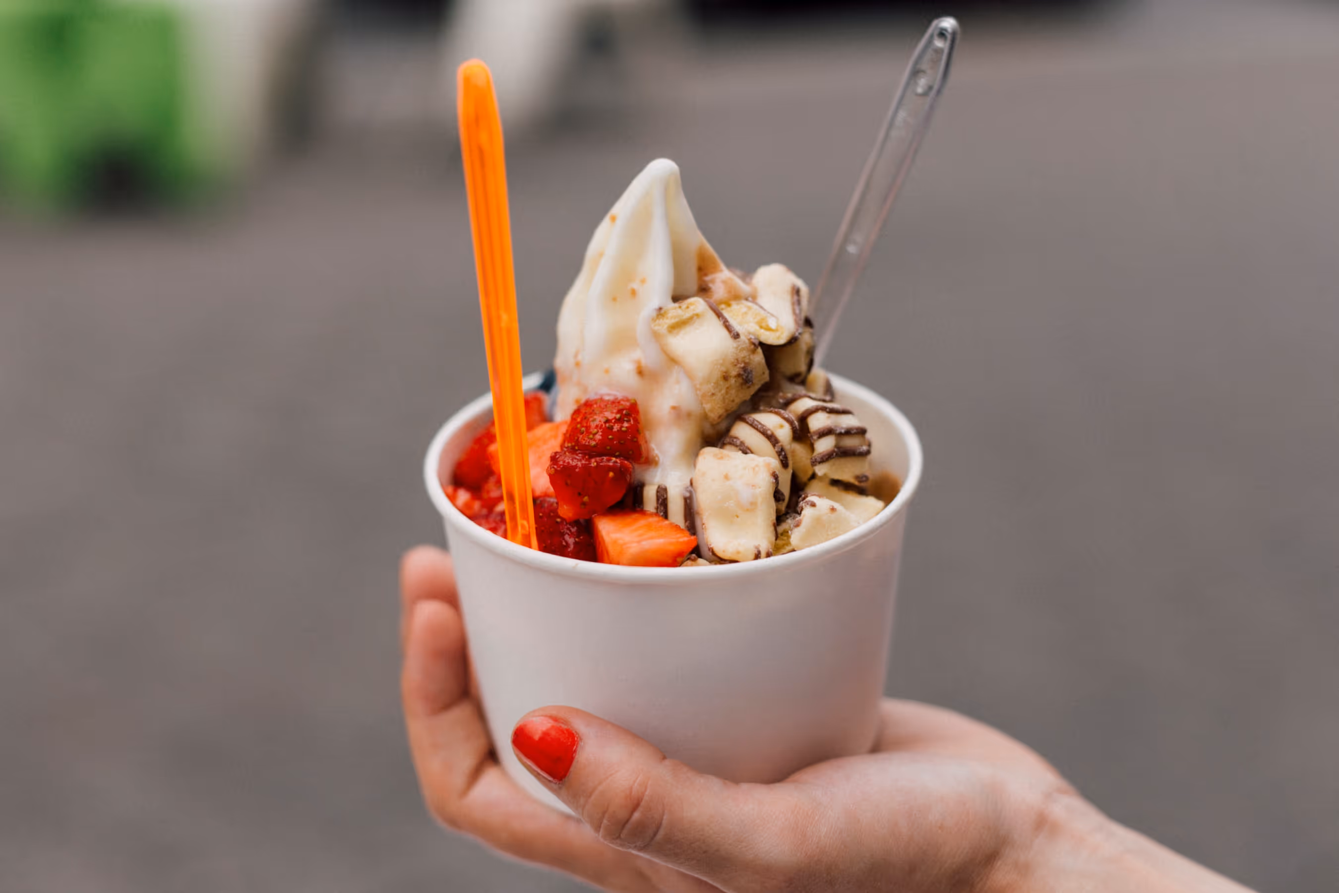 Hand holding a cup of soft-serve frozen yogurt topped with strawberries and chocolate-drizzled cereal pieces, with an orange spoon and a clear spoon.