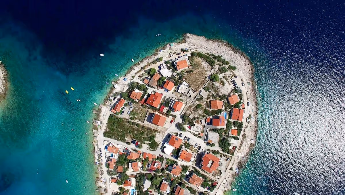 Aerial view of a small island with red-roofed houses surrounded by vibrant blue and turquoise sea.