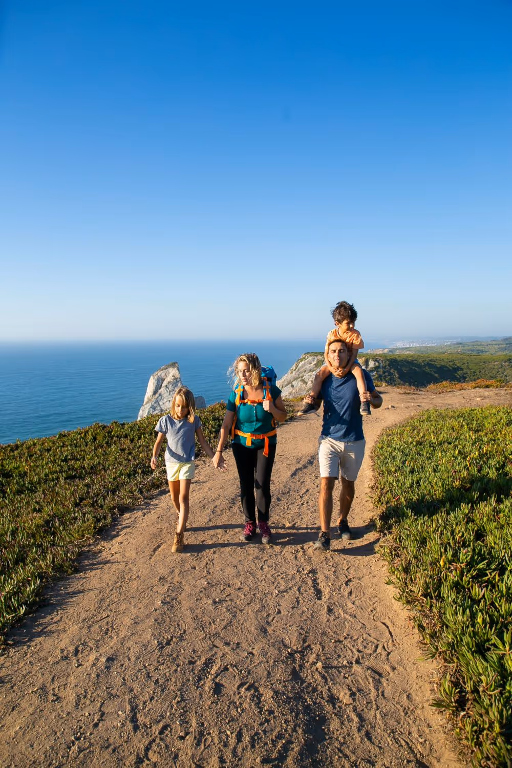 Family of four hiking on a coastal trail with ocean and cliffs in the background under a clear blue sky.