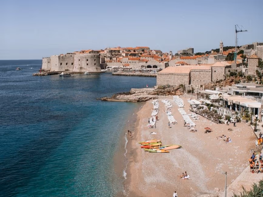 View of a coastal beach with rows of white umbrellas, kayaks on the sand, and historic stone buildings with red roofs in the background by the sea.