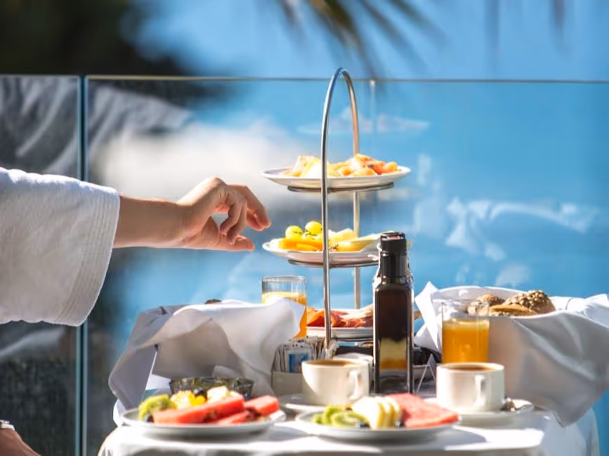 Hand reaching for fruit on a tiered serving tray with breakfast items, drinks, and coffee cups on a table with a glass railing and blue sky background.