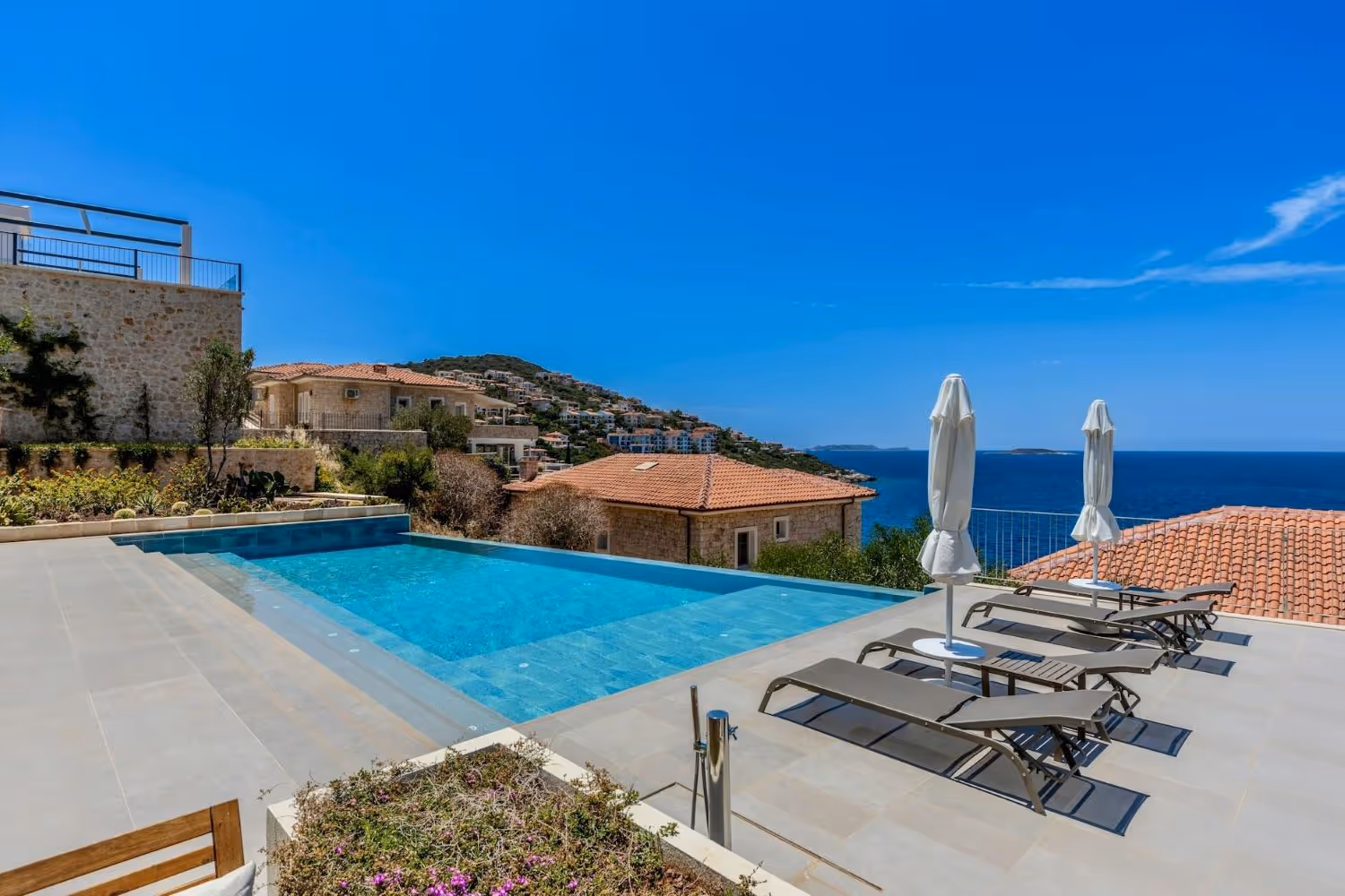 Infinity pool overlooking a sea view with sun loungers and closed umbrellas on a terrace beside stone houses with tiled roofs.