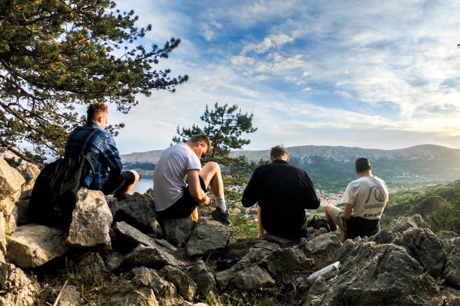 Four young men sitting on rocky hillside overlooking a valley and mountains under a partly cloudy sky.