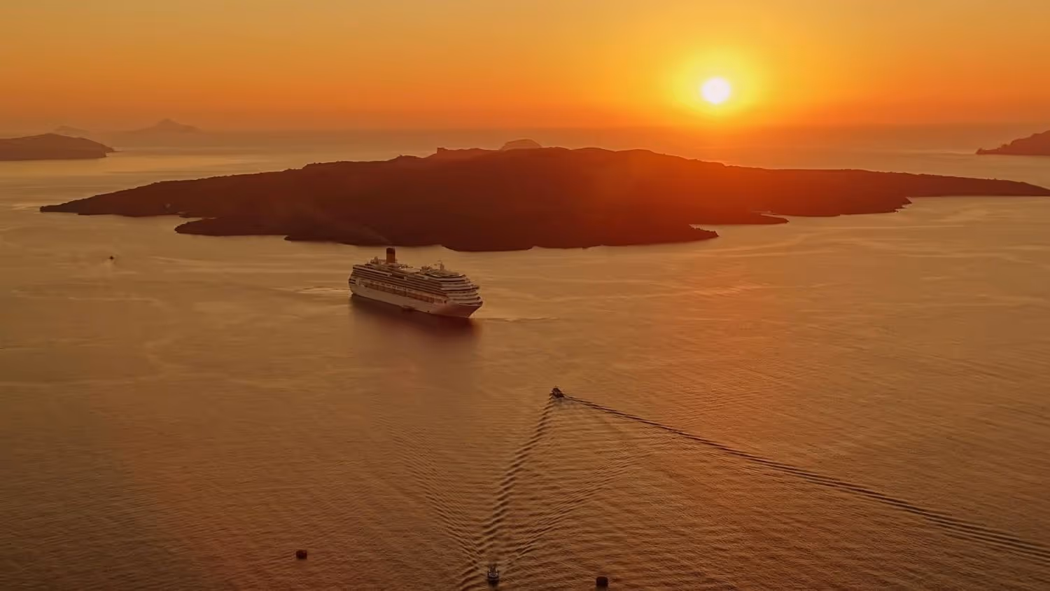 Cruise ship sailing near an island at sunset with orange sky and water reflections.