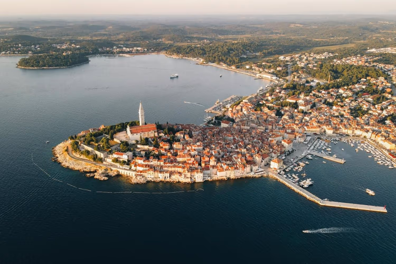 Aerial view of a coastal town with red-roofed buildings, a prominent church tower, surrounding harbor, and blue sea.