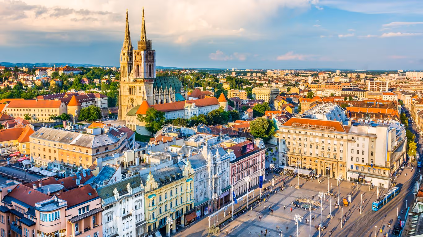 Aerial view of Zagreb city center featuring the Zagreb Cathedral with twin spires and surrounding historic buildings under a partly cloudy sky.