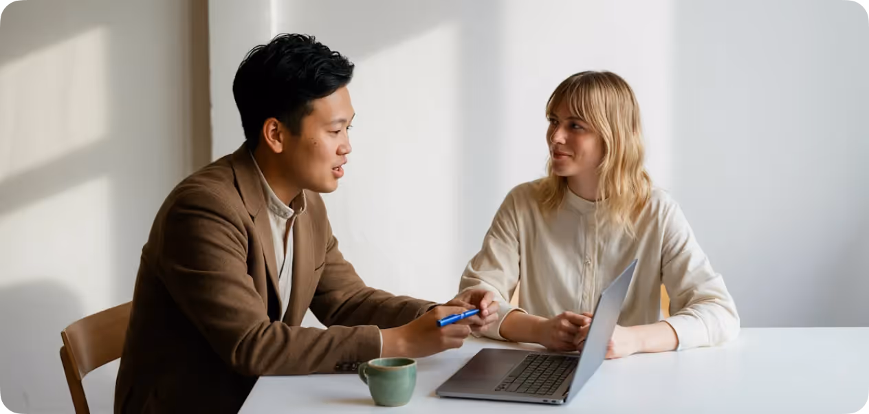 Two colleagues sitting at a table, discussing with a laptop and a coffee mug in a bright room.