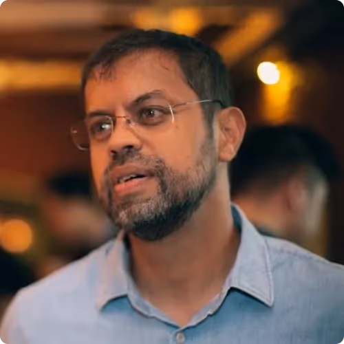 Man with glasses and a beard wearing a light blue shirt in a warmly lit indoor setting.
