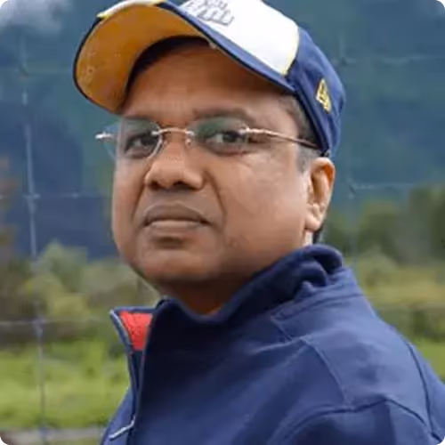 Man wearing glasses, a blue jacket, and a blue and white cap standing outdoors with blurred greenery in the background.