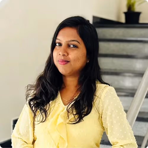 Woman with long black hair wearing a yellow blouse, standing indoors near stairs with a potted plant in the background.