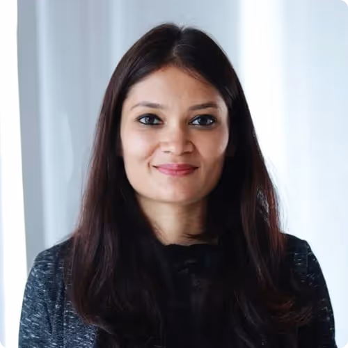 Portrait of a smiling woman with long dark hair standing against a light background.