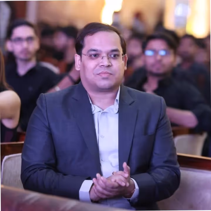 Man wearing glasses and a dark suit jacket clapping hands while seated at an event with other attendees blurred in the background.