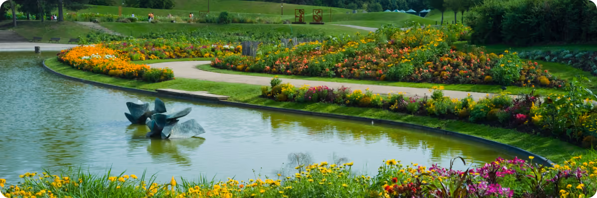 Curved pond with stone sculptures surrounded by vibrant flower beds in a green park.