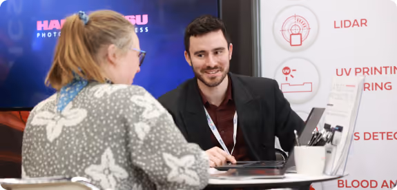Man and woman talking across a table with laptop at a technology booth with product icons.