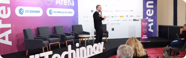 Speaker presenting on stage at Techinnox event with audience seated in front and purple branded backdrop.
