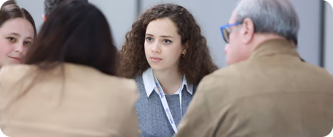 Young woman with curly hair and lanyard attentively listening to two people in a meeting.