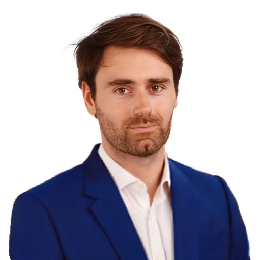 Young man with brown hair and beard in a blue suit jacket and white shirt against blurred city background.