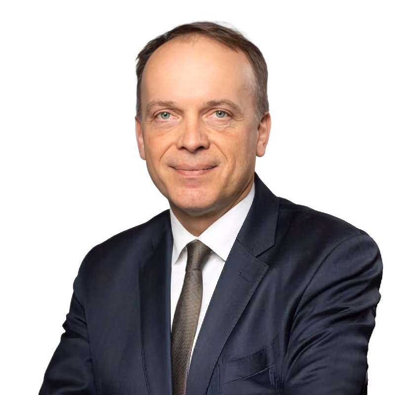 Smiling middle-aged man in a dark suit, white shirt, and tie against a plain gray background.