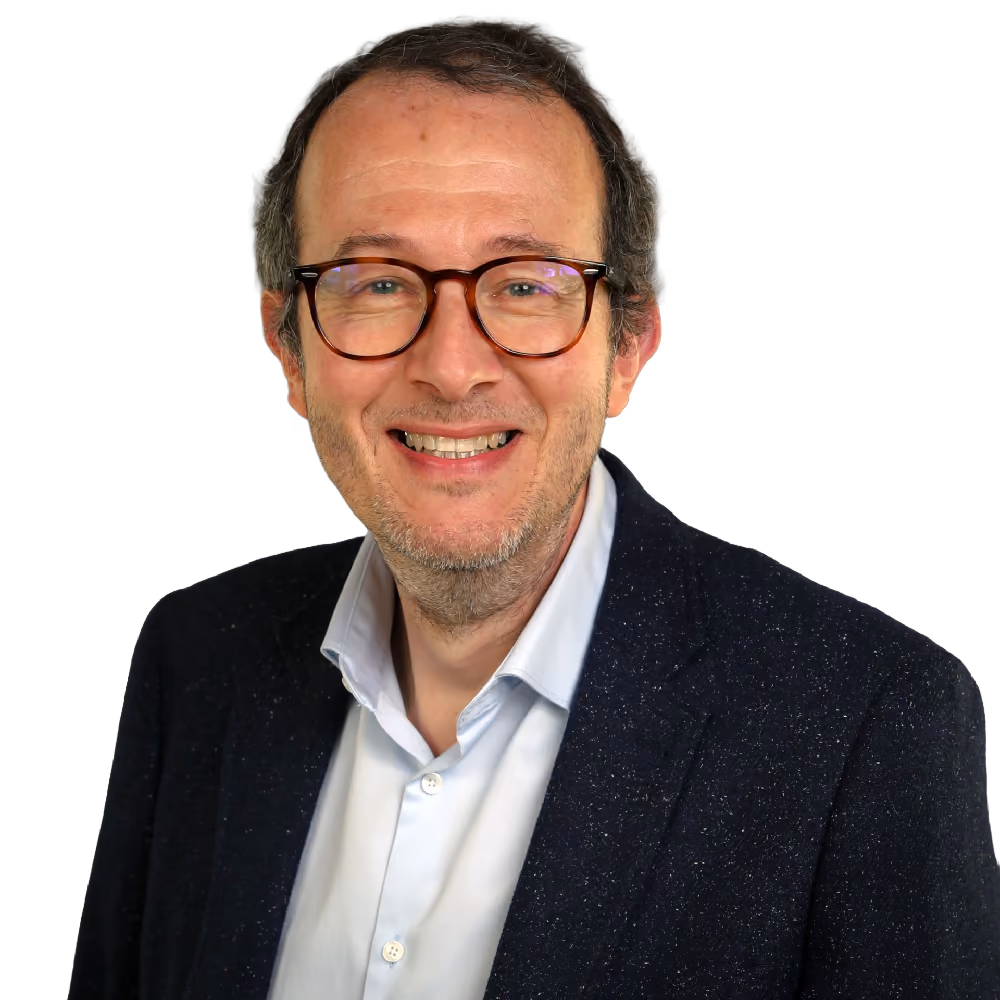 Smiling middle-aged man with glasses wearing a dark blazer and light blue shirt against gray background.