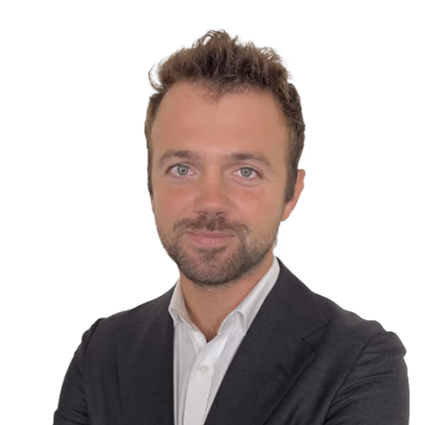 Smiling man with short curly hair and beard wearing a white shirt and dark blazer against white background.