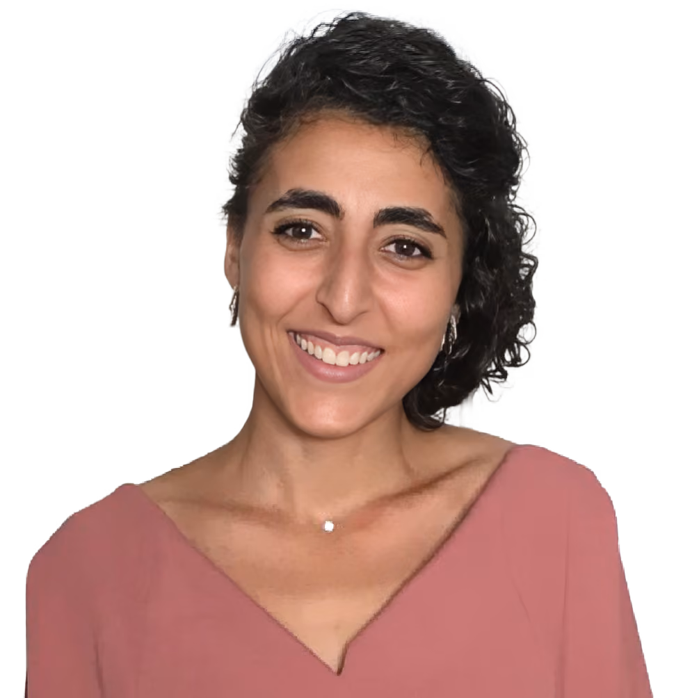 Smiling woman with curly dark hair wearing a rose-colored top and small earrings in front of a door.