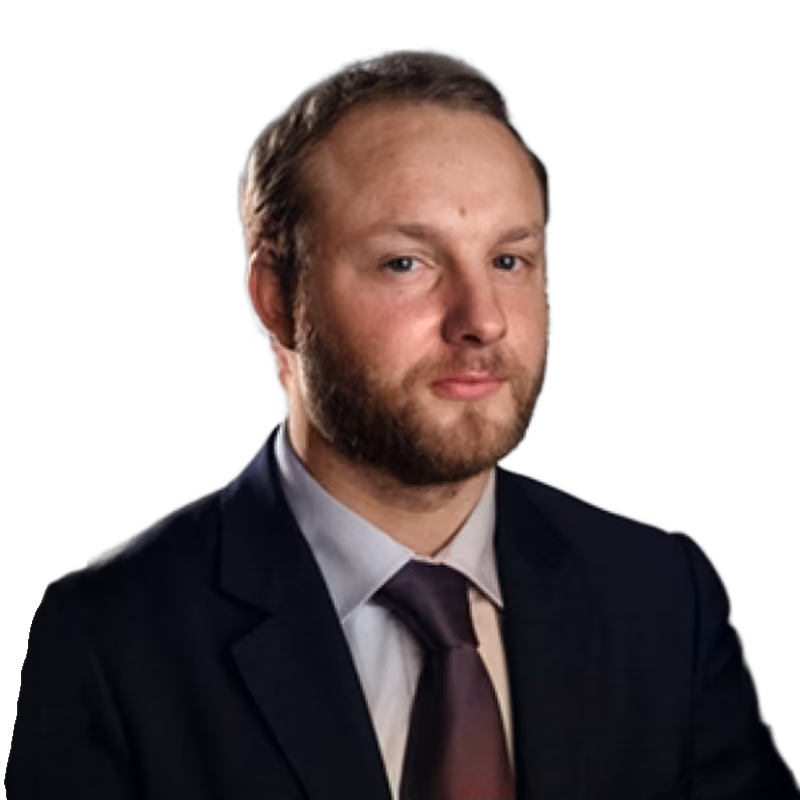 Portrait of a man with a beard wearing a dark suit, white shirt, and purple tie against a gray background.
