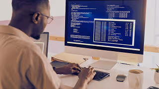 Man working at a desk with a keyboard and mouse, viewing multiple windows of code on a large computer monitor.