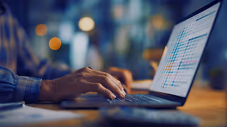 Person typing on a laptop keyboard with a spreadsheet displayed on the screen in a dimly lit workspace.