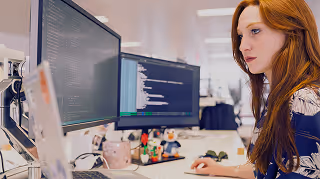 A woman with red hair focused on coding displayed on two computer monitors in an office.