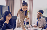 Three professionals in a meeting, with one woman standing and pointing at documents on a table while two colleagues listen attentively.
