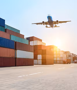 Airplane flying above stacked shipping containers at a port during sunset.
