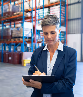 Woman in business attire writing on a clipboard inside a warehouse with shelves of boxes in the background.