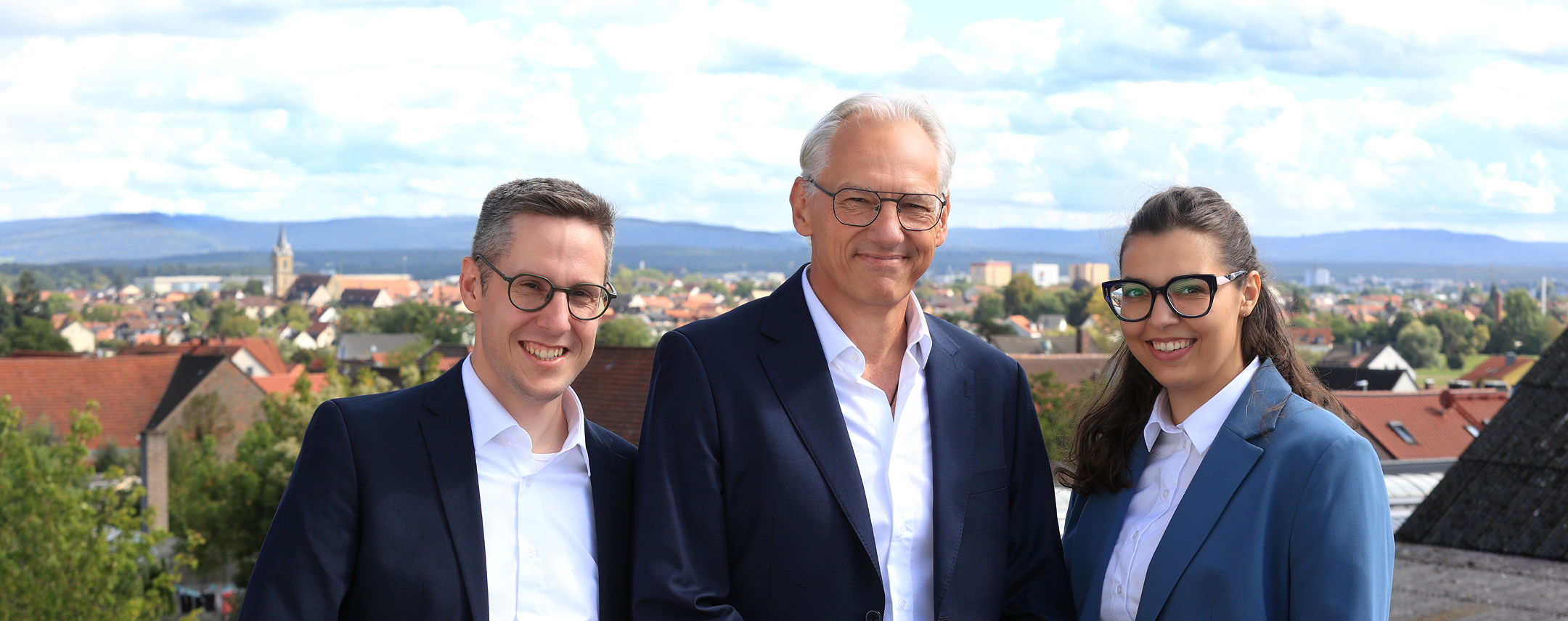 Three smiling professionals wearing glasses and suits stand outdoors with a town and hills in the background.