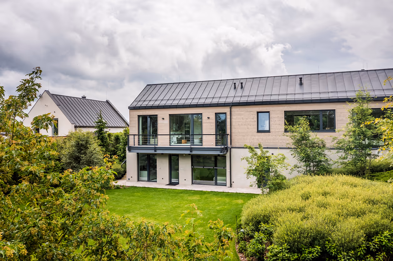 Moderní dvoupodlažní dům s balkonem, velkými okny a zelenou zahradou za oblačného počasí.