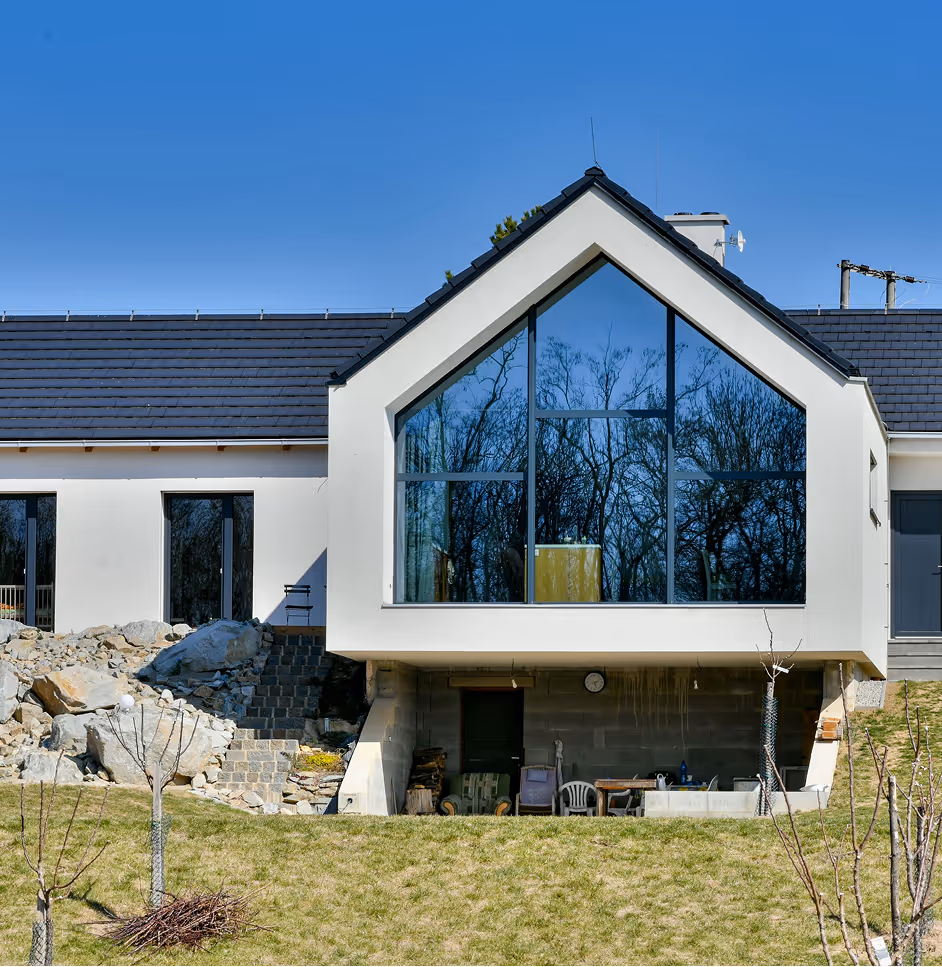 Moderní dům s velkým trojúhelníkovým oknem odrážejícím stromy a travnatým předzahrádkou.