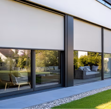 Moderní dům s velkými okny, venkovními žaluziemi a posezením na terase s výhledem do zahrady.