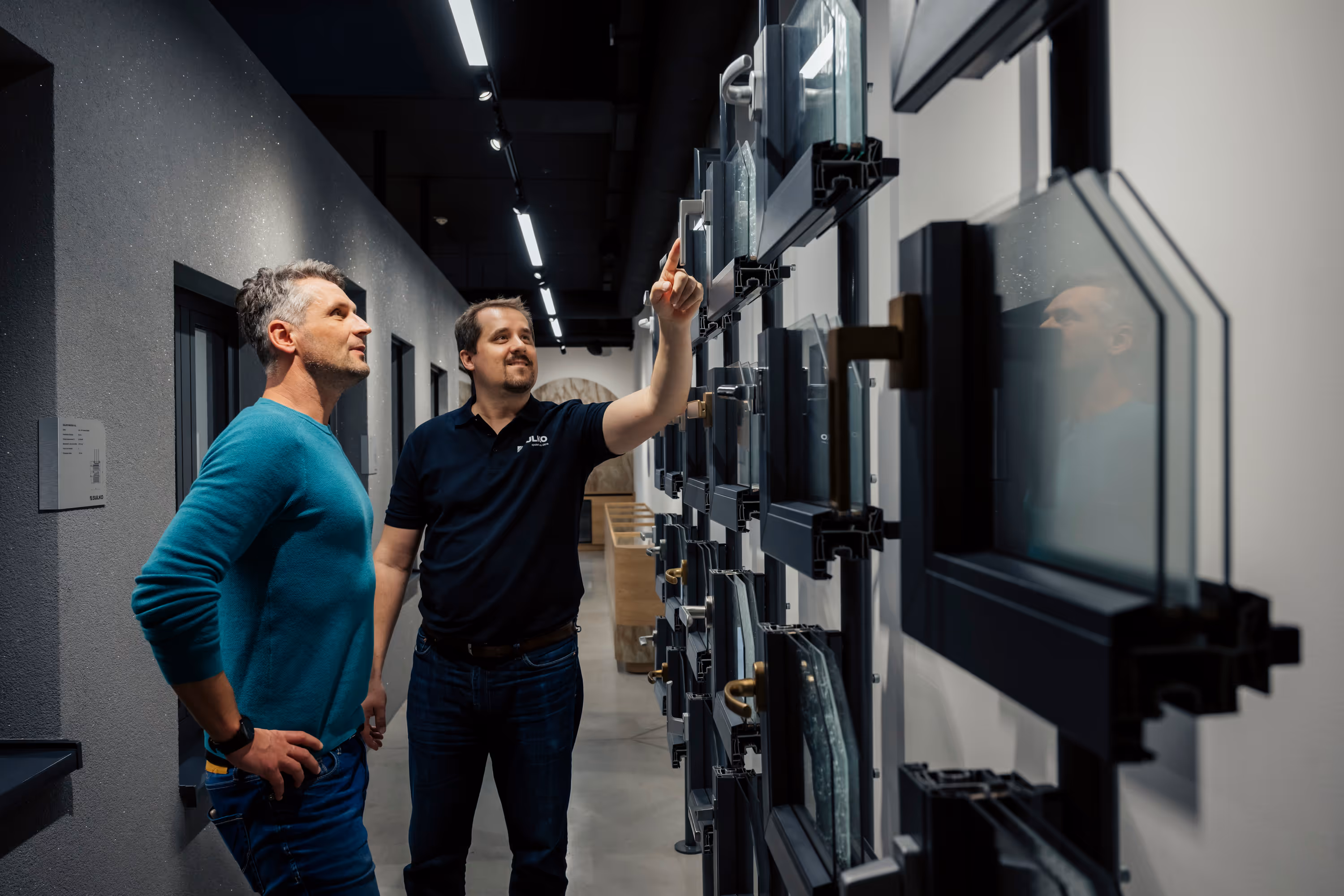 Dva muži diskutují a prohlížejí nástěnný panel s různými vzorky okenních rámů a skel v moderní místnosti.