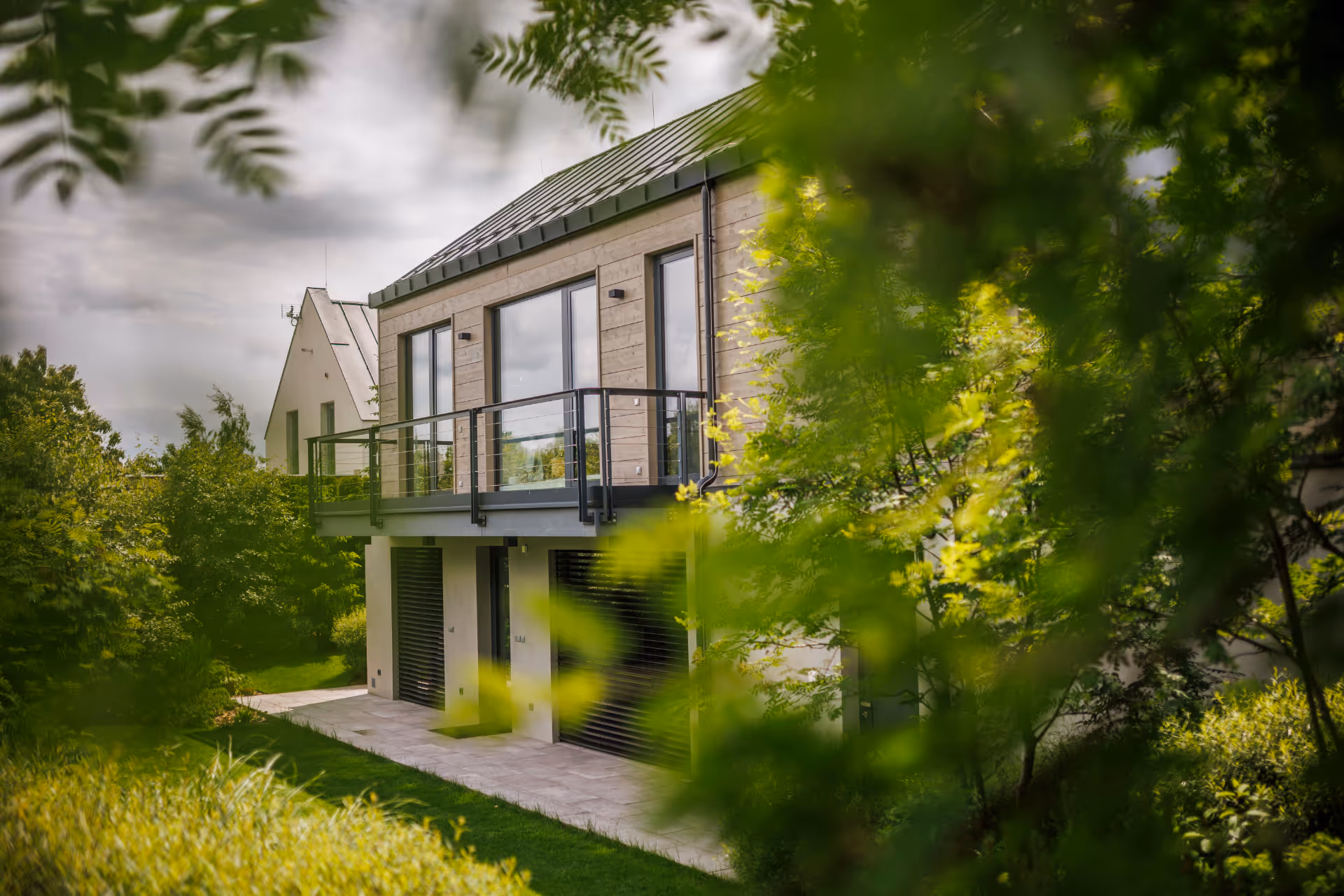 Moderní dům se skleněným balkonem, částečně zakrytý zelenými stromy a keři.