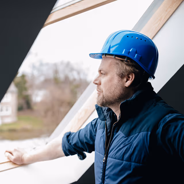 Muž v modré ochrané přilbě a tmavé vestě, který stojí u okna ve stavbě a dívá se ven.