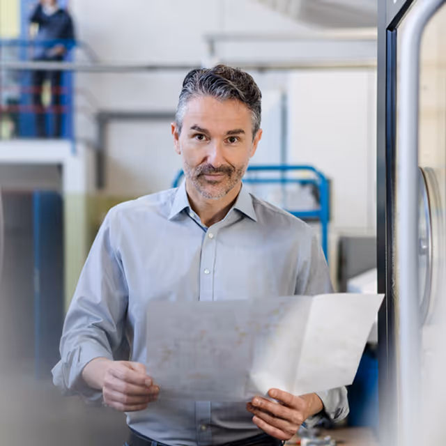 Muž v šedé košili drží papírový plán v průmyslovém prostředí.