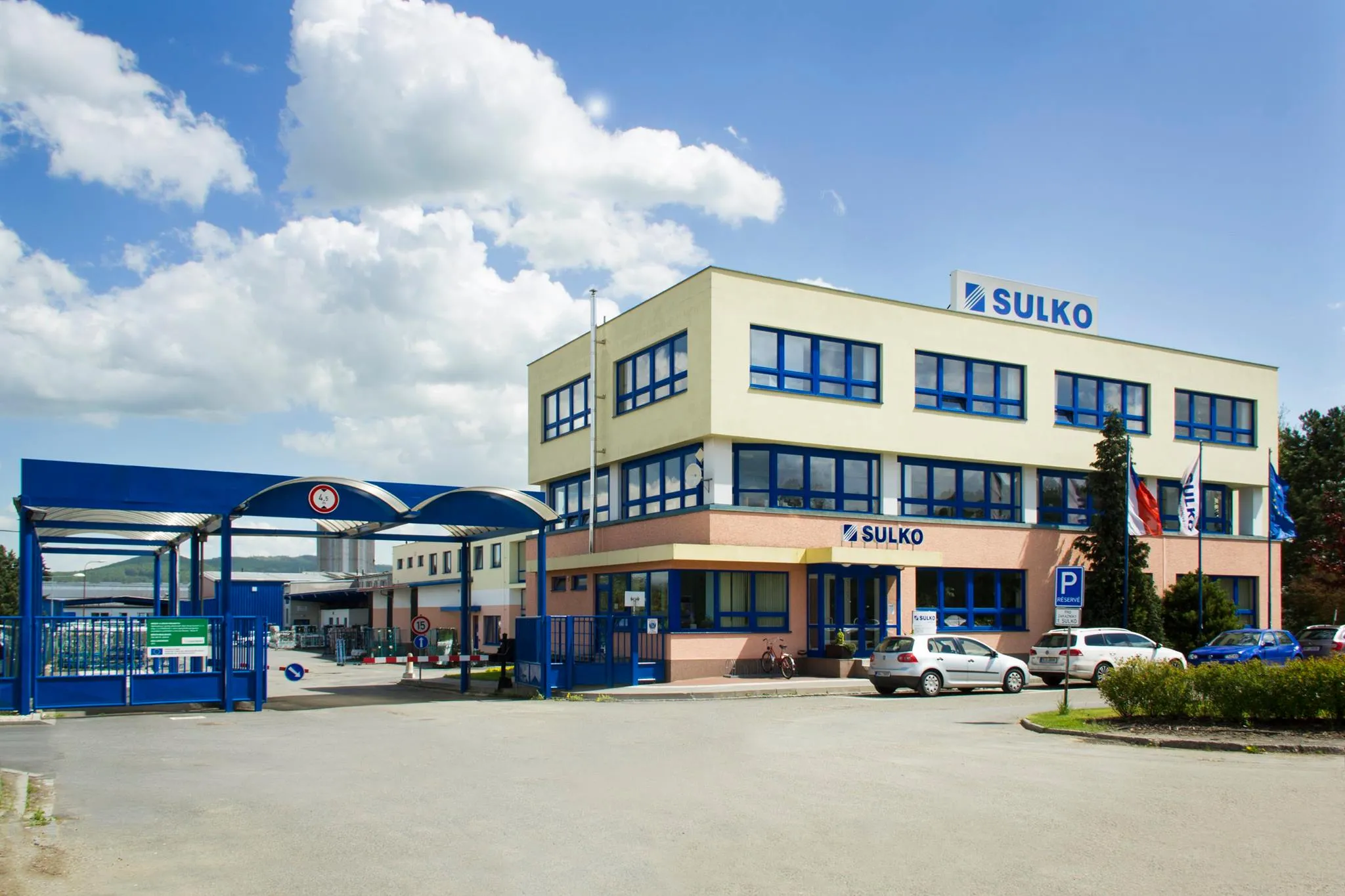 Budova společnosti SULKO s parkovištěm a vjezdem s modrou bránou pod oblačnou oblohou.