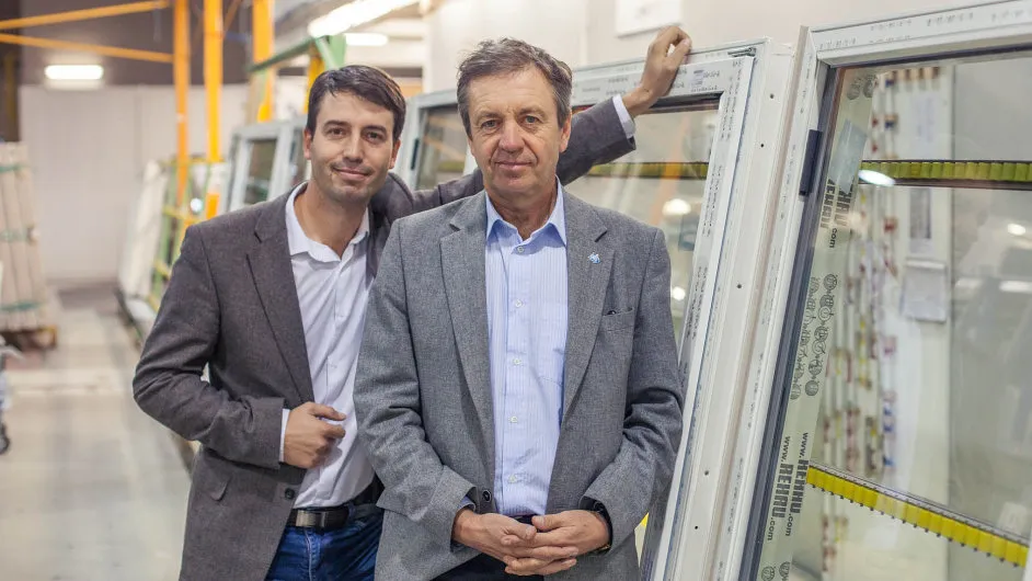 Dva muži v saku stojí vedle okenních rámů v průmyslovém prostředí.