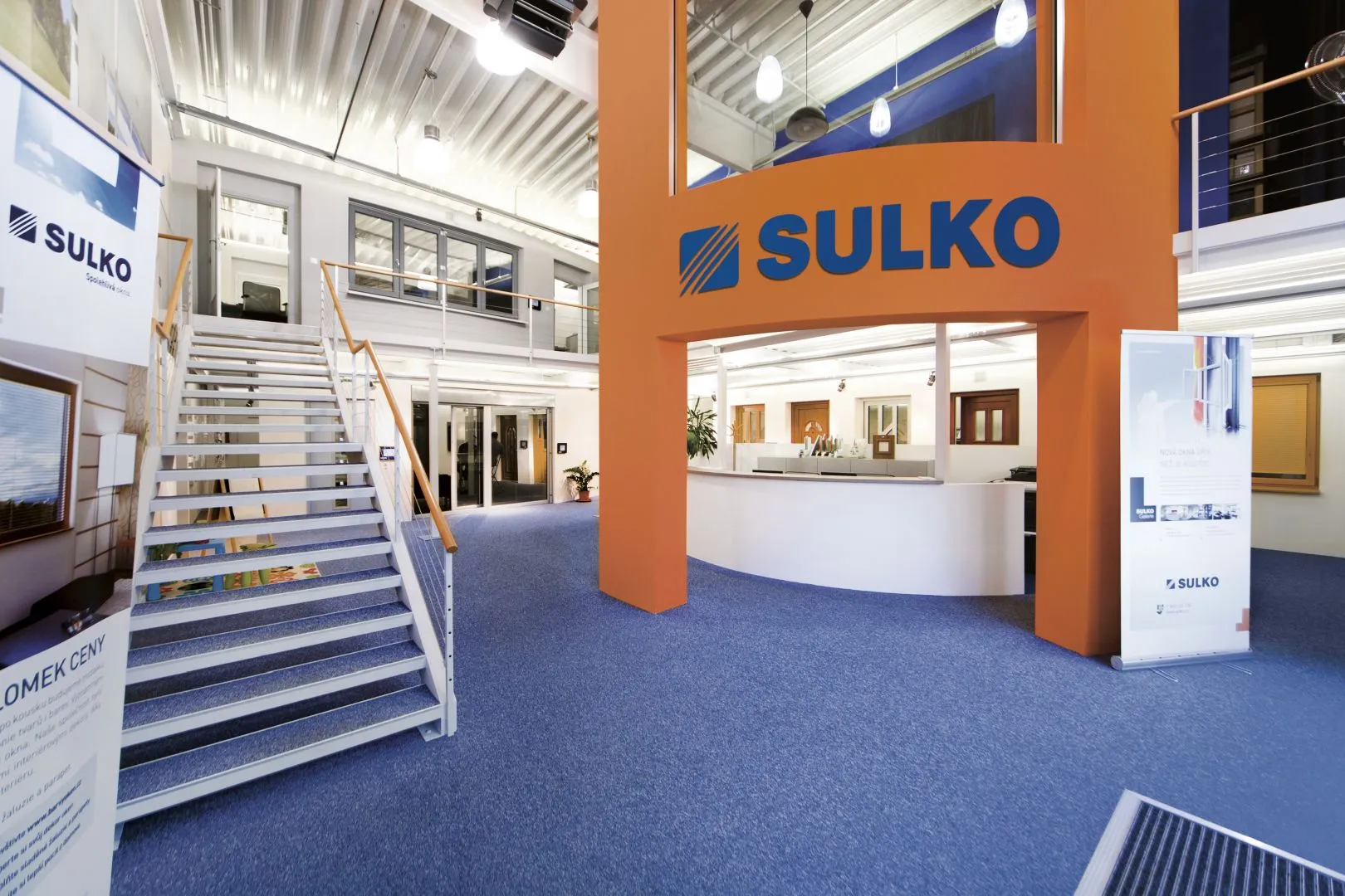 Moderní kancelářská vstupní hala s modrým kobercem, schodištěm a oranžovým sloupem s nápisem SULKO.